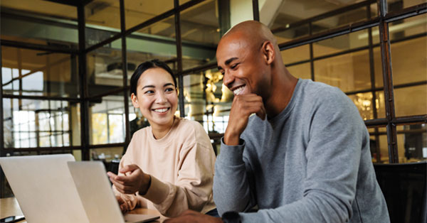 Two colleagues smiling while working together on laptops in a modern office with large glass windows in the background.