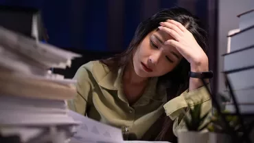 A stressed woman in a green shirt with a hand on her head in a dark office with piles of papers and books on either side of her