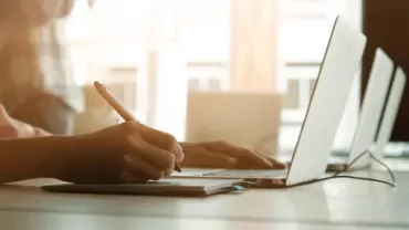 Person writing on a tablet next to a laptop.