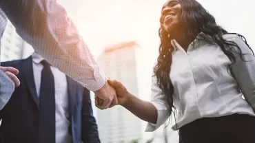 Two professionals shaking hands outdoors with a skyscraper in the background.